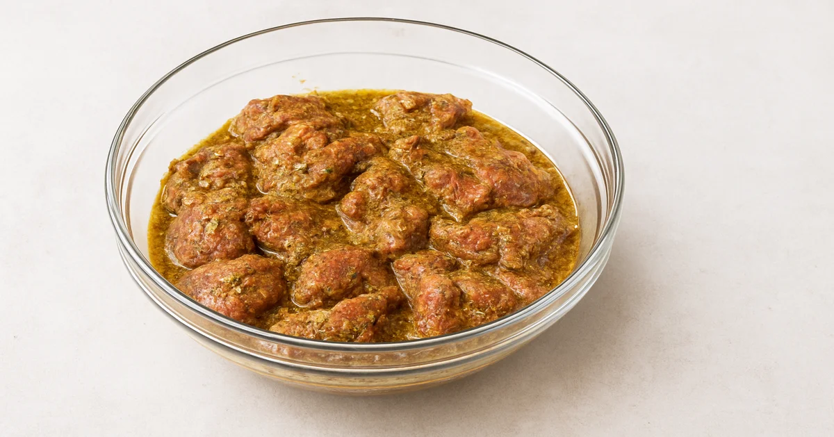 Chunks of lamb marinating in a spiced herb mixture inside a clear glass bowl on a light surface